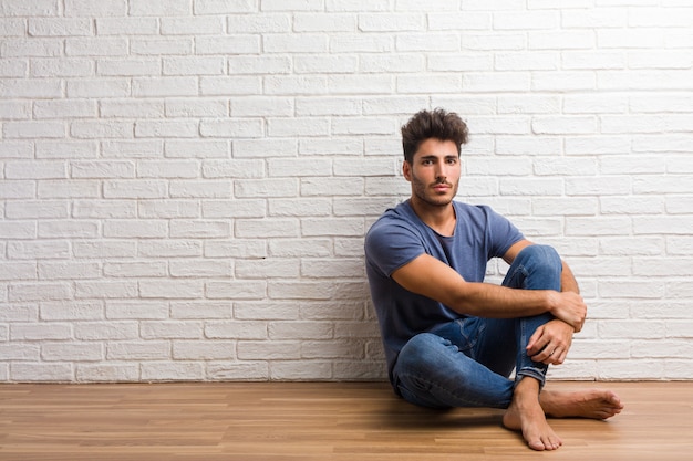 El hombre natural joven se sienta en un piso de madera cruzando sus brazos, sonriendo y feliz, siendo confiado y amigable