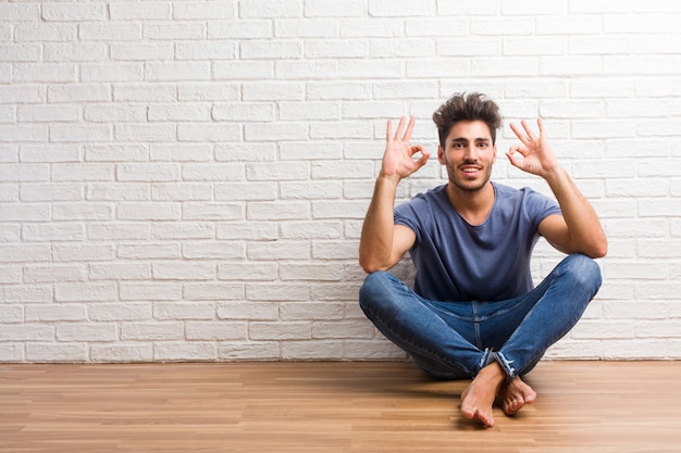 El hombre natural joven se sienta en un suelo de madera alegre y confiado que hace gesto aceptable