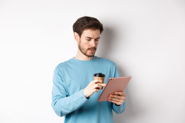 Hombre navegando en línea en tableta digital y bebiendo café de la taza de papel, de pie sobre fondo blanco. Copia espacio