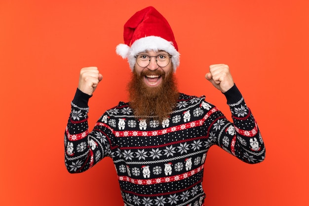 Hombre de Navidad con barba larga sobre pared roja aislada celebrando una victoria
