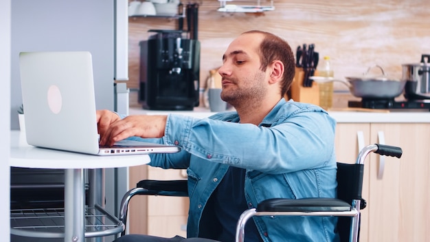 Hombre con necesidades especiales en silla de ruedas trabajando en equipo portátil en la cocina. Hombre corporativo con parálisis minusvalía discapacidad minusválidos dificultades trabajando después de un accidente teniendo internet videollamada en línea co