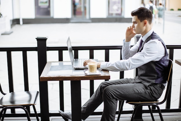 Hombre de negocios acertado joven que se sienta en café urbano al aire libre con el ordenador portátil y que habla en el teléfono