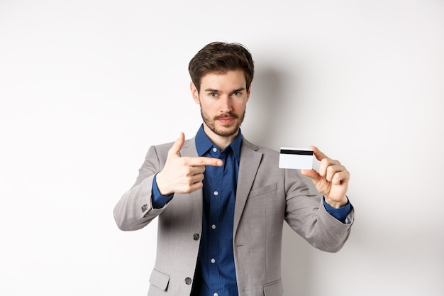 Hombre de negocios acertado en traje apuntando a la tarjeta de crédito plástica y sonriendo, recomendando el banco, de pie sobre fondo blanco.