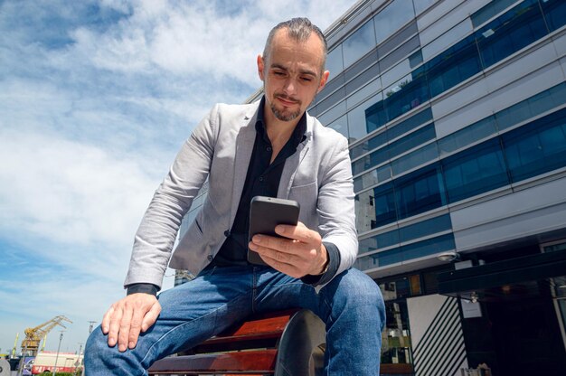 Hombre de negocios adulto caucásico francés con barba vestido con ropa elegante sentado al aire libre con edificios comerciales alrededor usando el teléfono enviando mensajes espacio para copiar