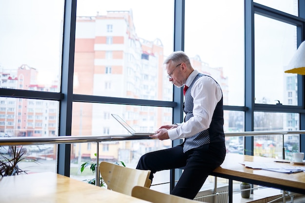 Hombre de negocios adulto, profesor, mentor trabajando en un nuevo proyecto. Se sienta junto a una gran ventana sobre la mesa. Mira la pantalla del portátil.