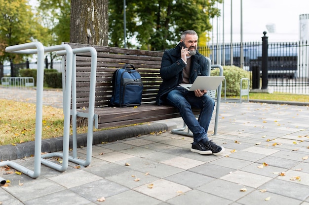 Hombre de negocios adulto que trabaja en línea en una computadora portátil y llama por teléfono en el parque de la ciudad