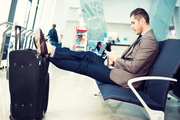 Hombre de negocios en el aeropuerto con teléfono inteligente y maleta revisando correos electrónicos antes de abordar