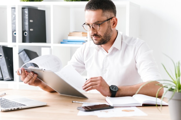 Hombre de negocios sin afeitar concentrado de 30 años en camisa blanca sosteniendo y mirando el portapapeles con documentos en papel, durante el trabajo en la oficina