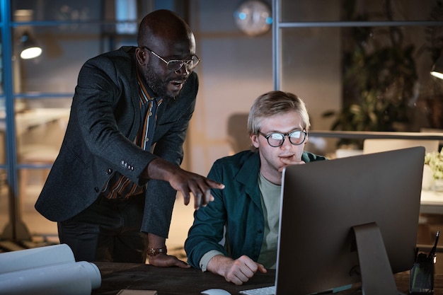 Hombre de negocios africano apuntando al monitor de la computadora y discutiendo el proyecto en línea con su colega mientras trabajan en la oficina durante la fecha límite