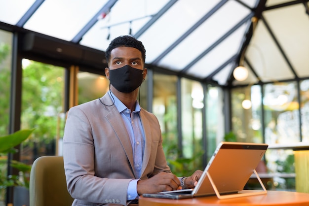 Hombre de negocios africano en la cafetería con ordenador portátil y vistiendo mascarilla