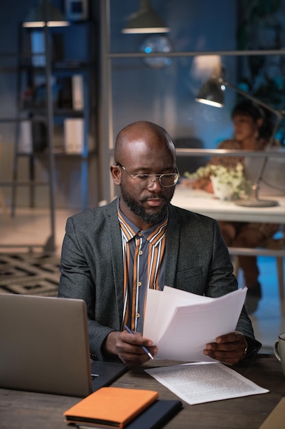 Hombre de negocios africano concentrándose en su papeleo, sentado en la mesa frente a la computadora portátil y leyendo documentos durante su trabajo en la oficina hasta tarde