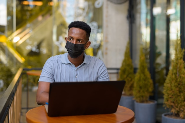 Hombre de negocios africano con máscara facial y distanciamiento social mientras está sentado en la cafetería usando una computadora portátil