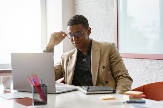 Un hombre de negocios africano ocupado con gafas trabajando en una laptop mientras está sentado en un coworking moderno