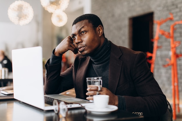 Hombre de negocios africano que usa el teléfono y que bebe café mientras que trabaja en la computadora portátil en un restaurante.