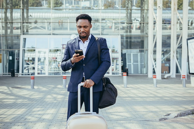 Hombre de negocios africano que viaja cerca del aeropuerto con maleta llamando a un taxi usando la aplicación móvil