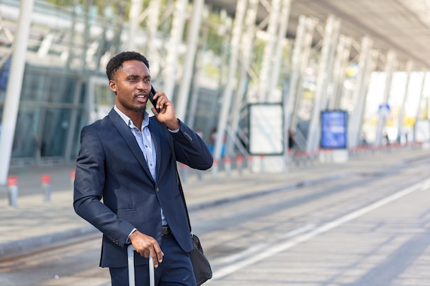 Hombre de negocios africano que viaja cerca del aeropuerto con maleta llamando a un taxi usando la aplicación móvil