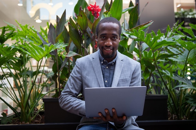 Un hombre de negocios africano sentado en un banco trabaja con su laptop. Persona exitosa con sonrisas de mirada confidente. Empresario trabaja al aire libre