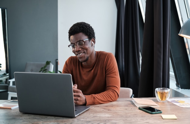 Hombre de negocios africano sentado en la mesa hablando en línea y sonriendo él usando laptop