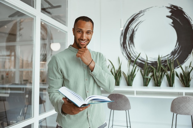 Un hombre de negocios africano sonriente parado en una oficina moderna y sosteniendo un libro