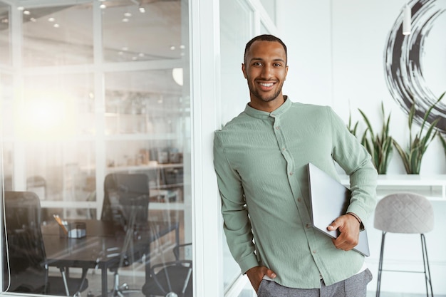 Hombre de negocios africano sonriente sosteniendo una computadora portátil mientras está de pie en una oficina acogedora y mirando la cámara