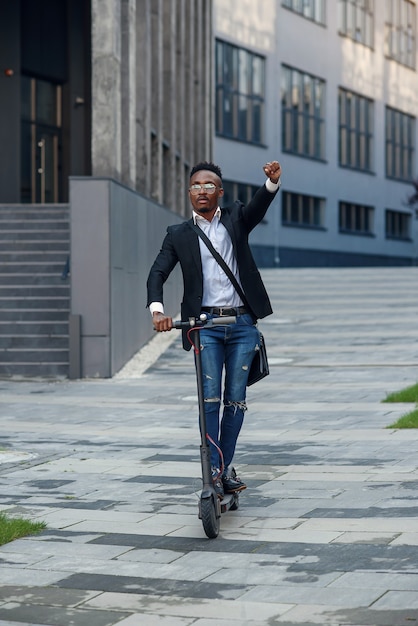 Hombre de negocios afroamericano alegre moderno monta scooter eléctrico después de terminar su jornada laboral