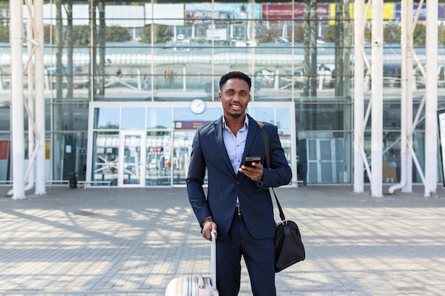 Hombre de negocios afroamericano confiado joven acertado que se mueve con el teléfono móvil en las manos contra el fondo del edificio de oficinas moderno El hombre de negocios negro céntrico camina usando un teléfono móvil en traje formal