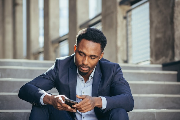 Hombre de negocios afroamericano deprimido leyendo malas noticias desde el teléfono celular, frustrado y triste sentado en las escaleras en traje de negocios