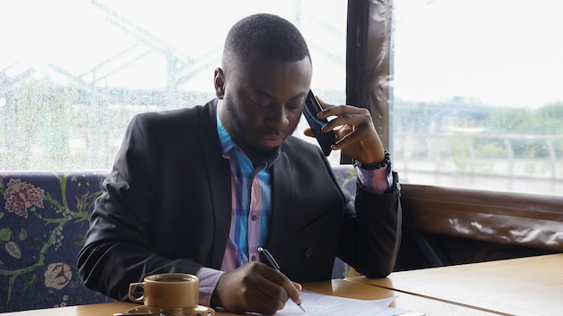 Hombre de negocios afroamericano está llamando al teléfono inteligente y tomando café en la cafetería.