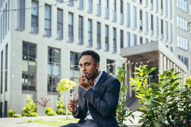 Foto hombre de negocios afroamericano fumando cannabis al aire libre sentado en un banco del parque de la ciudad en el fondo de la calle urbana. hombre de negocios empleado de sexo masculino. oficinista en traje alivia el estrés de una marihuana en el exterior