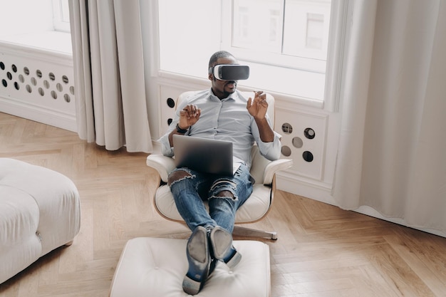 Hombre de negocios afroamericano con gafas VR trabaja en realidad virtual sentado en un sillón con una computadora portátil