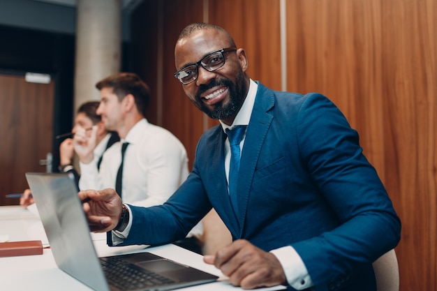 Hombre de negocios afroamericano negro sentado a la mesa con el portátil en el fondo de la gente del equipo de oficina.