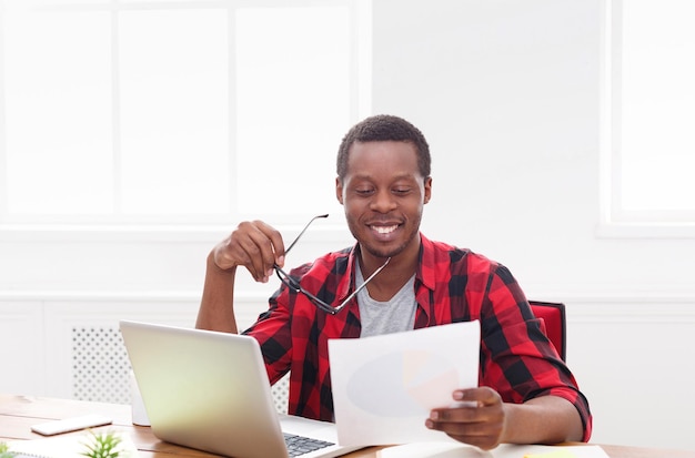 Hombre de negocios afroamericano en la oficina, trabajar con ordenador portátil y documentos con placer, espacio de copia