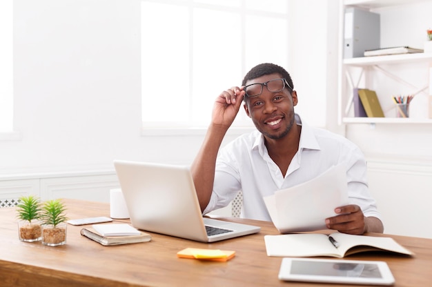 Hombre de negocios afroamericano en la oficina, trabajar con ordenador portátil y documentos con placer, mirando a cámara, espacio de copia