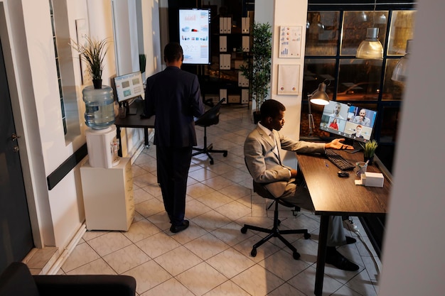 Hombre de negocios afroamericano que tiene una discusión remota con el equipo durante una conferencia de reunión de videollamada en línea, discutiendo la estrategia de marketing. Diversos compañeros de trabajo que trabajan tarde en la noche en la oficina de inicio