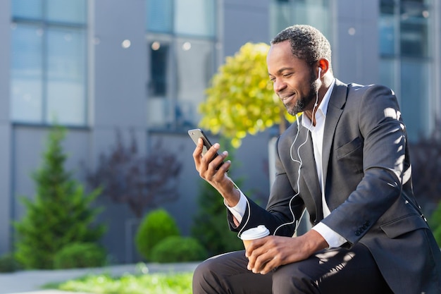 Hombre de negocios afroamericano que usa el teléfono sentado en un banco fuera del edificio de oficinas mirando al jefe