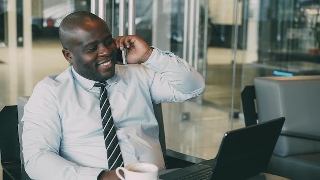 Hombre de negocios afroamericano con ropa formal hablando en su teléfono inteligente y navegando por la red mientras mira su computadora portátil en un café moderno durante el almuerzo
