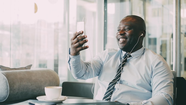 Hombre de negocios afroamericano con ropa formal riéndose y hablando con su familia a través de un chat de video de un teléfono inteligente en un café aireado Él agita su mano alegremente