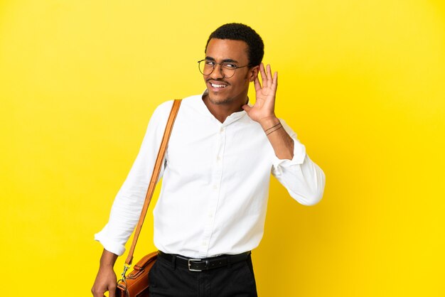 Hombre de negocios afroamericano sobre fondo amarillo aislado escuchando algo poniendo la mano en la oreja