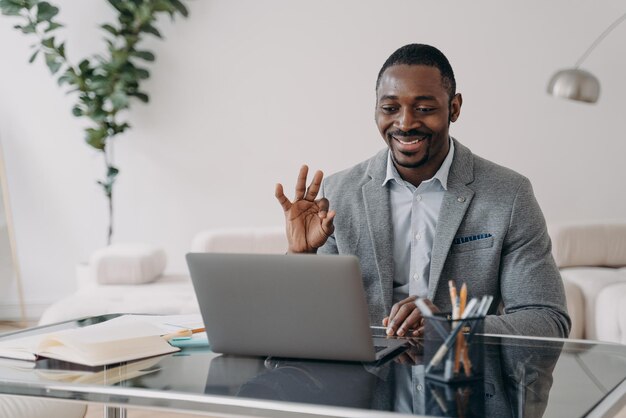 Un hombre de negocios afroamericano sonriente muestra un gesto correcto hablando por videollamada trabajando en una computadora portátil