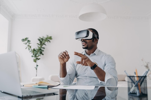 El hombre de negocios afroamericano usa gafas de realidad virtual trabajando en una computadora portátil en el escritorio de la oficina Hightech