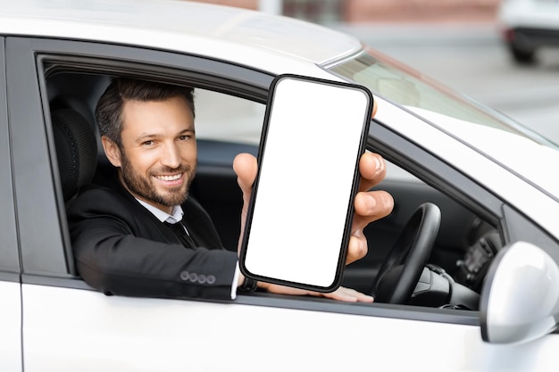 Hombre de negocios alegre sentado en un auto mostrando una maqueta de teléfono celular