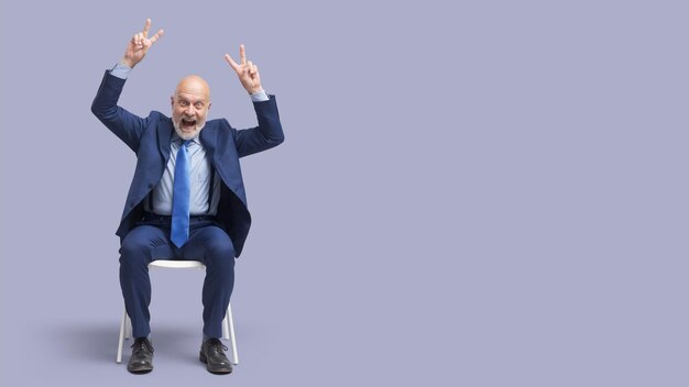 Foto un hombre de negocios alegre sentado en una silla y haciendo signos de v está celebrando su éxito