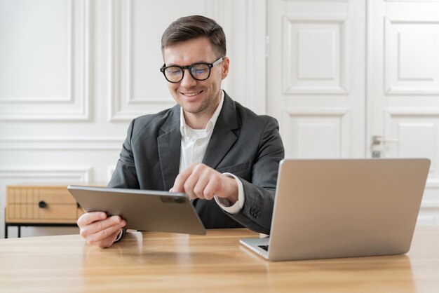 Un hombre de negocios alegre en un traje trabaja en una tableta con una computadora portátil abierta en el escritorio en la oficina