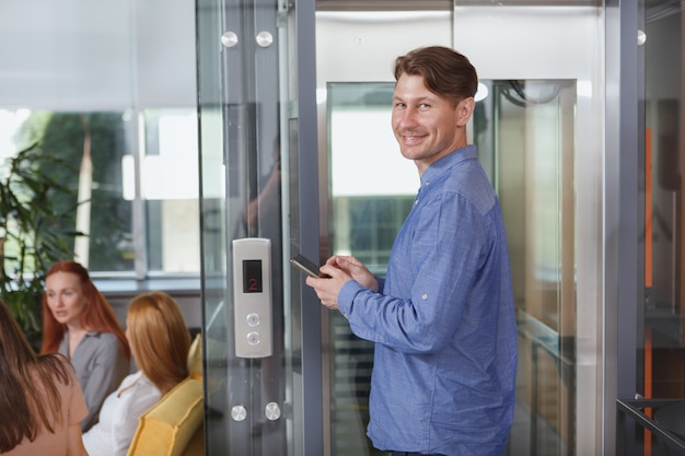 Hombre de negocios alegre usando un teléfono inteligente mientras espera un ascensor en el edificio de oficinas