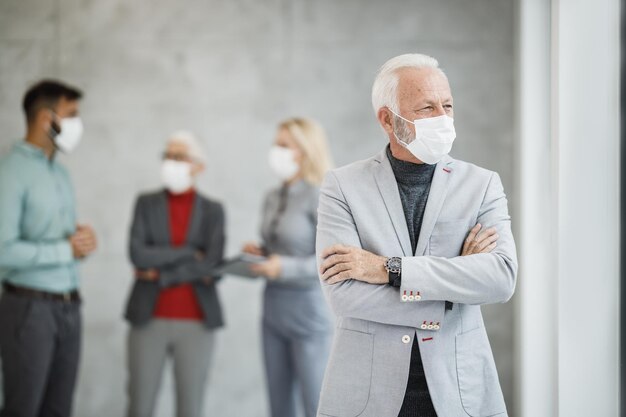 Un hombre de negocios de alto nivel confiado con máscara protectora parado en una oficina moderna y mirando por la ventana durante la pandemia del virus de la corona.