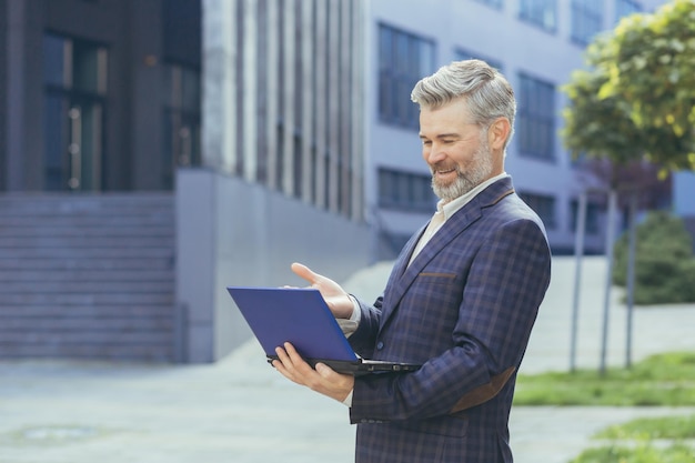 Un hombre de negocios de alto rango se para cerca de un edificio de oficinas y sostiene una netbook en sus manos habla