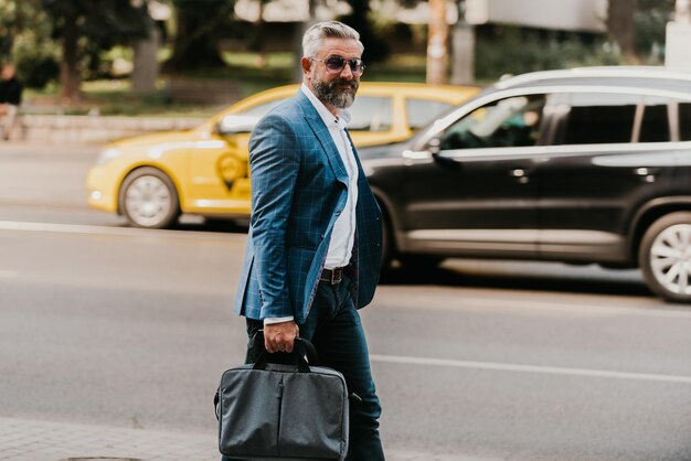 Un hombre de negocios de alto rango con traje y gafas de sol caminando por la ciudad mientras lleva una bolsa.