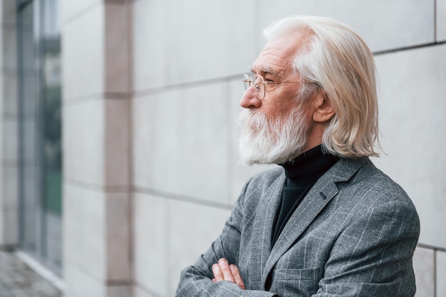 Un hombre de negocios de alto rango vestido con ropa formal con cabello gris y barba está al aire libre parado contra la pared