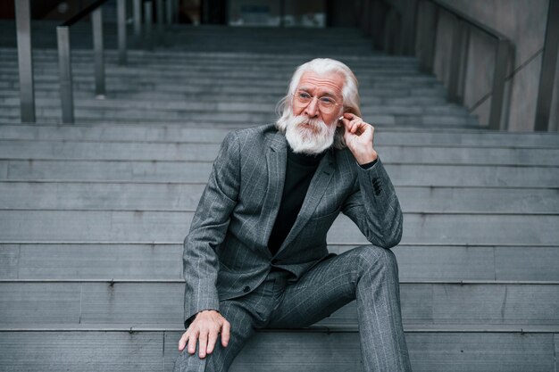 Un hombre de negocios de alto rango vestido con ropa formal con cabello gris y barba está al aire libre sentado en las escaleras