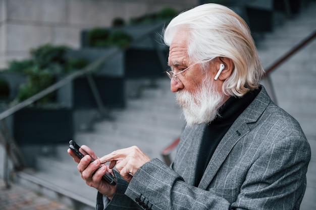 Un hombre de negocios de alto rango vestido con ropa formal con pelo gris y barba está al aire libre con auriculares inalámbricos y teléfono